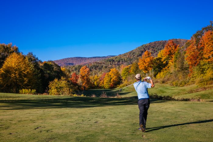 A golfer follows through on a swing on a course surrounded by vibrant fall foliage and rolling hills.