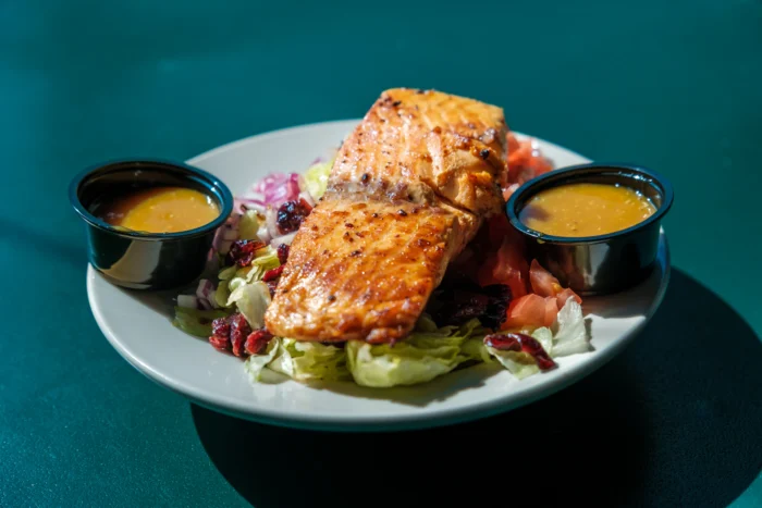 Grilled salmon fillet served on a white plate with fresh lettuce, red onions, cranberries and two small black ramekins of yellow sauce against a teal background