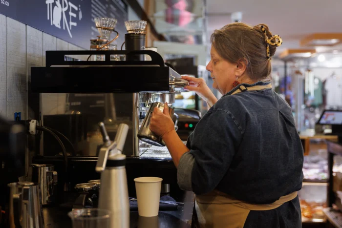 A woman standing in front of an espresso machine, steaming milk in a cup. 