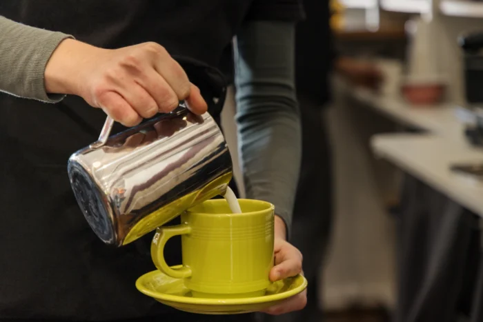 A person pouring steamed milk from a silver pot into a green mug, resting on a green saucer.