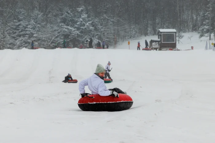 A person in a light purple winter jacket and green hat rides down a snowy slope on a red and black snow tube. In the background, other people are tubing and there's a small wooden structure near snow-covered trees.