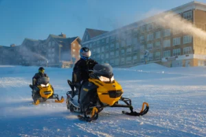 Two snowmobilers riding yellow Ski-Doo snowmobiles across a snowy field at a ski resort. The riders wear black protective gear and helmets. A large lodge-style building with multiple stories is visible in the background, and snow machines are creating artificial snow.