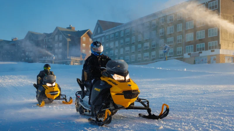 Two snowmobilers riding yellow Ski-Doo snowmobiles across a snowy field at a ski resort. The riders wear black protective gear and helmets. A large lodge-style building with multiple stories is visible in the background, and snow machines are creating artificial snow.