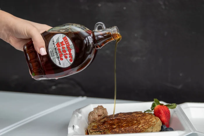 A hand pouring maple syrup from a decorative glass bottle onto a pancake garnished with a strawberry and blueberries on a white plate.