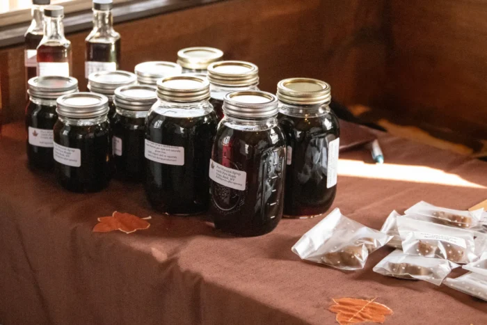 Multiple mason jars filled with dark maple syrup arranged in a row on a brown tablecloth with maple leaf decorations. Small bags, possibly containing maple products, are visible in the foreground.