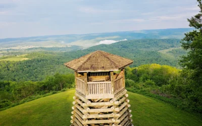 A wooden observation tower with a hexagonal top sits on a green hilltop, overlooking a lush landscape of forested mountains and rolling hills with distant fields.