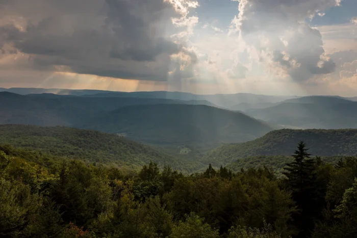 A mountain landscape with multiple rolling forested hills receding into the distance. Sunbeams pierce through clouds. The foreground shows dense forest with deciduous and evergreen trees, while the background features layers of mountains fading into a misty blue haze.