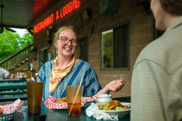 An outdoor dining scene at a restaurant with a "Restaurant & Lodging" neon sign visible. A woman with blonde hair, glasses, and wearing a blue striped outfit with a yellow patterned scarf is smiling at someone across the table. There are drinks and food on the table, with the woman appearing to be mid-conversation in a casual dining setting with red checkered food baskets.