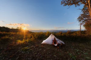 Person sits beside a small tent reading during sunset on a mountain overlook with distant hills