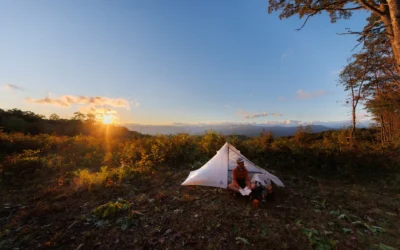 Person sits beside a small tent reading during sunset on a mountain overlook with distant hills