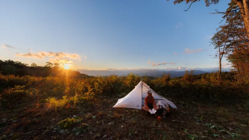 Person sits beside a small tent reading during sunset on a mountain overlook with distant hills