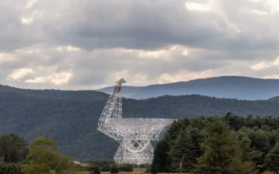 A large radio telescope with a white metal dish structure set against a backdrop of green forested mountains and a cloudy sky.