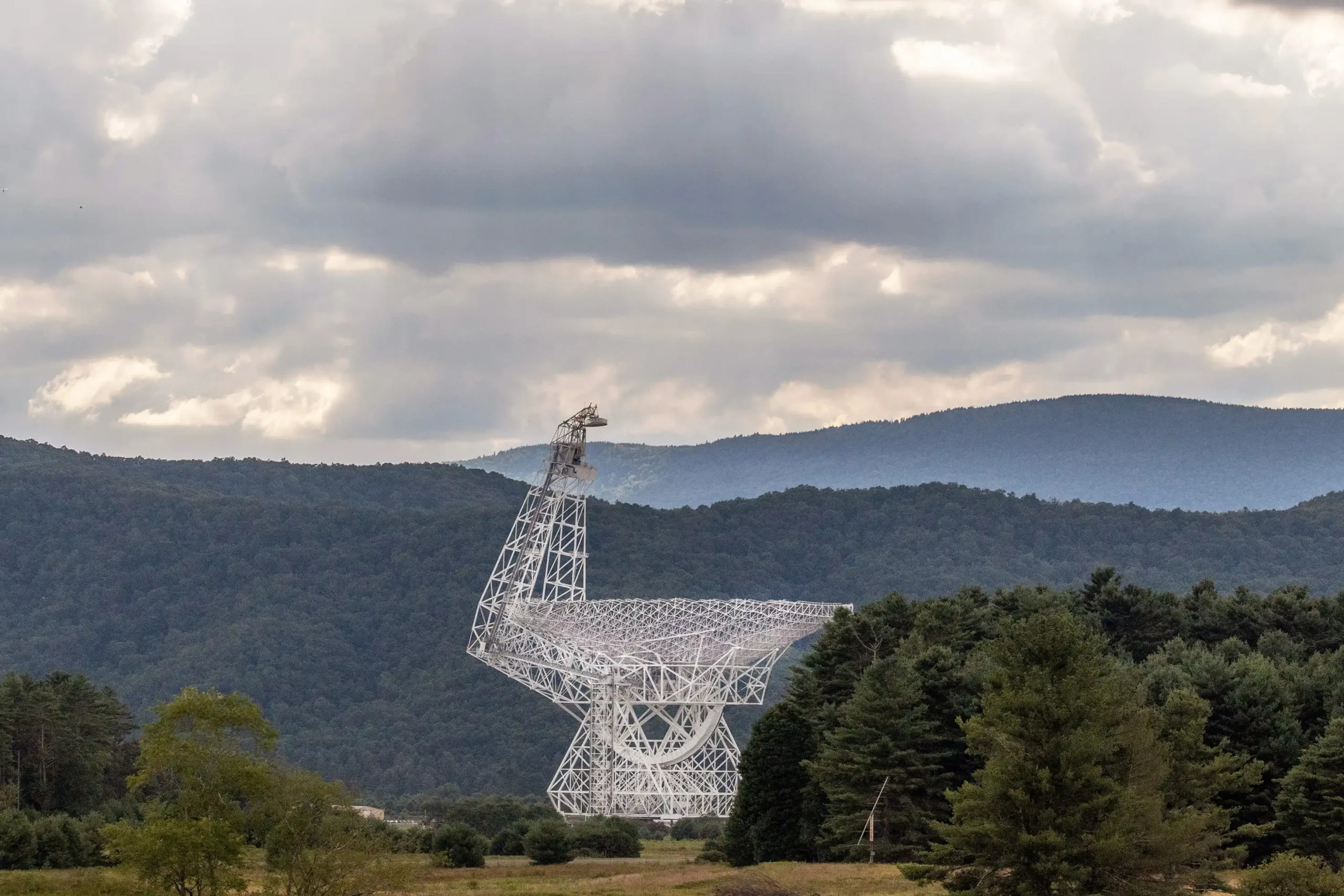 A large radio telescope with a white metal dish structure set against a backdrop of green forested mountains and a cloudy sky.