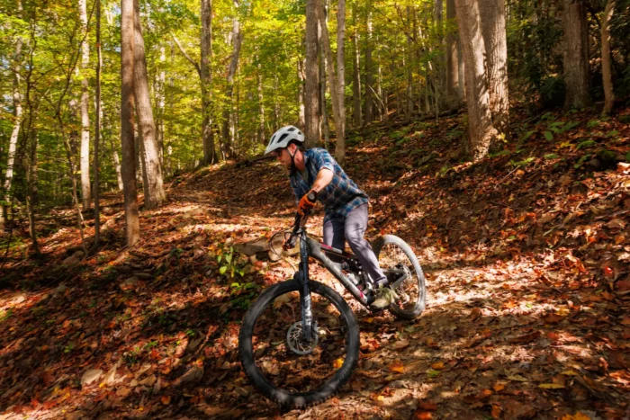 A mountain biker riding a trail through a forest in autumn, navigating over fallen leaves and roots.