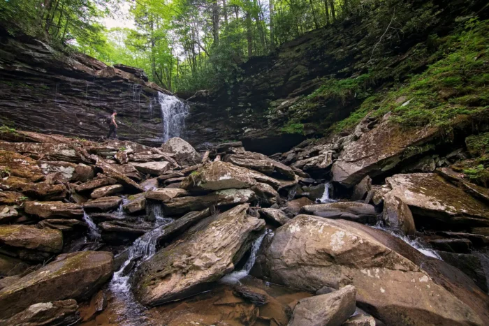 A rocky waterfall flows through a boulder-filled gorge with a person visible on the rocks, surrounded by lush forest.