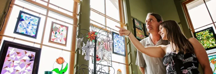 Couple admiring colorful stained-glass artwork displayed in a brightly lit gallery with large wooden-framed windows.