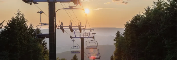 Ski lift chairs suspended above a grassy mountain slope at sunset, with evergreen trees framing the view and distant ridgelines.
