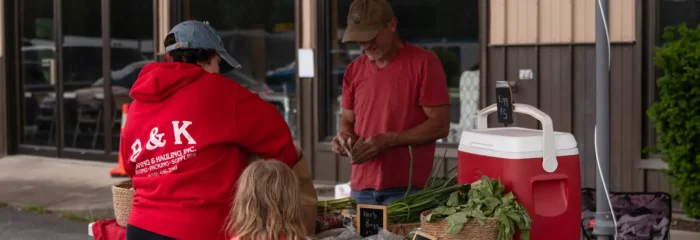 Local vendor selling fresh vegetables at an outdoor farmers market stand, interacting with a woman and child under a red canopy.