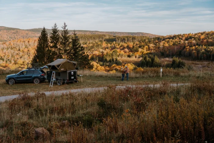 SUV with rooftop tent and two people standing in a field with autumn trees and hills