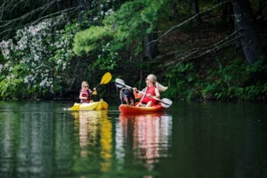 A woman and child in separate kayaks on a calm lake, with a black dog standing on the woman's orange kayak. Both are wearing life jackets and sunglasses, surrounded by green hills and trees.