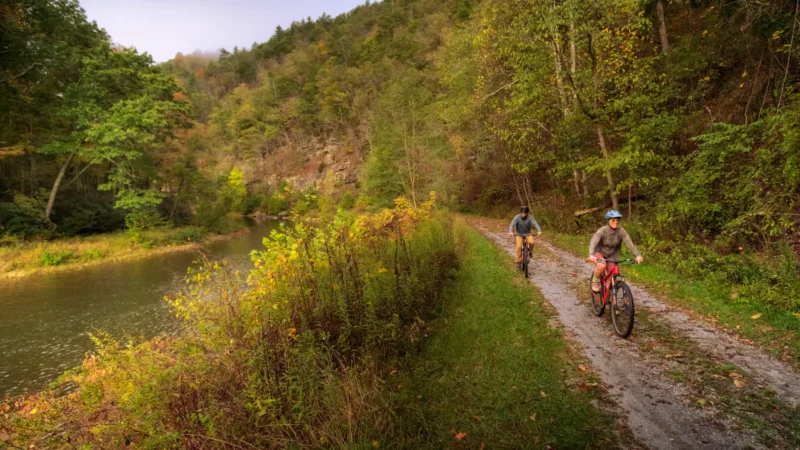 Two cyclists riding on a dirt path alongside a river, surrounded by hills and autumn foliage.