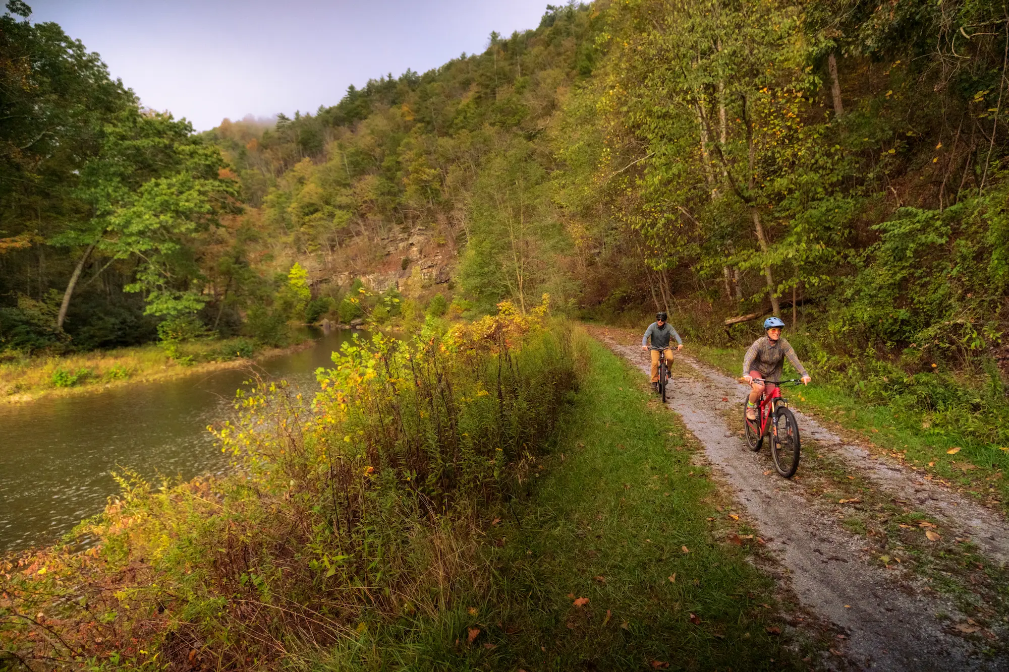 Two cyclists riding on a dirt path alongside a river, surrounded by hills and autumn foliage.