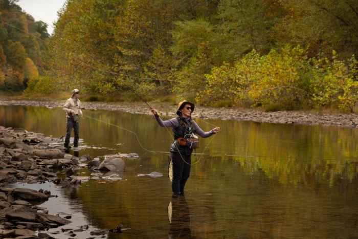 Two people fly fishing in a calm river, standing in waders with fishing rods extended, surrounded by trees displaying fall foliage along the rocky shoreline.