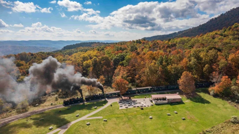 Steam train with black smoke traveling through autumn forest landscape with colorful fall foliage