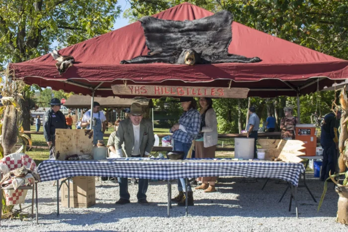 Market vendor in hat sitting at booth with "YHS Hillbillies" sign under red tent canopy