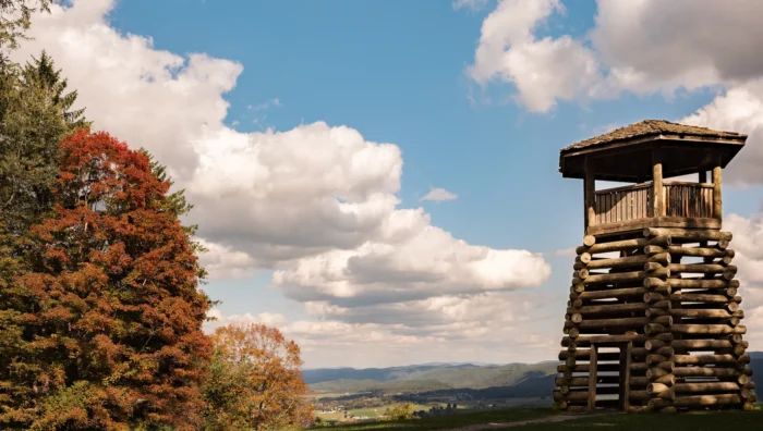 A wooden observation tower overlooks a valley landscape with autumn trees and distant mountains under a blue sky with white clouds.