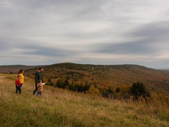 A family of four stands in a grassy field overlooking rolling hills covered in autumn foliage under an overcast sky.