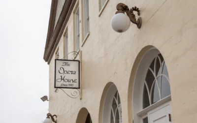 Exterior view of The Opera House building showing a cream-colored wall with arched windows, an ornate wall lamp, and a hanging sign that reads "The Opera House Circa 1910"