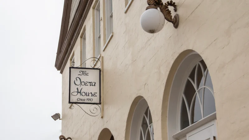 Exterior view of The Opera House building showing a cream-colored wall with arched windows, an ornate wall lamp, and a hanging sign that reads "The Opera House Circa 1910"