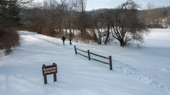 Two people walking on a snowy trail past a Greenbrier River Trail sign
