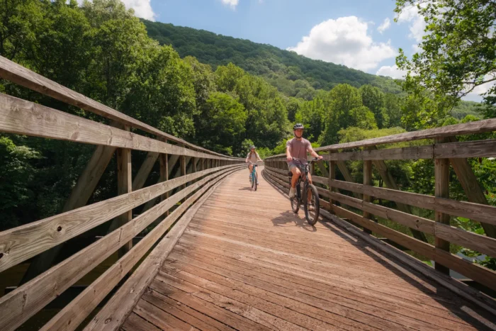 Two cyclists riding bikes across a wooden trestle bridge surrounded by lush green hills.