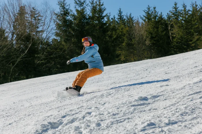 Snowboarder wearing a light blue jacket and brown pants, carving down a snowy slope on a sunny day