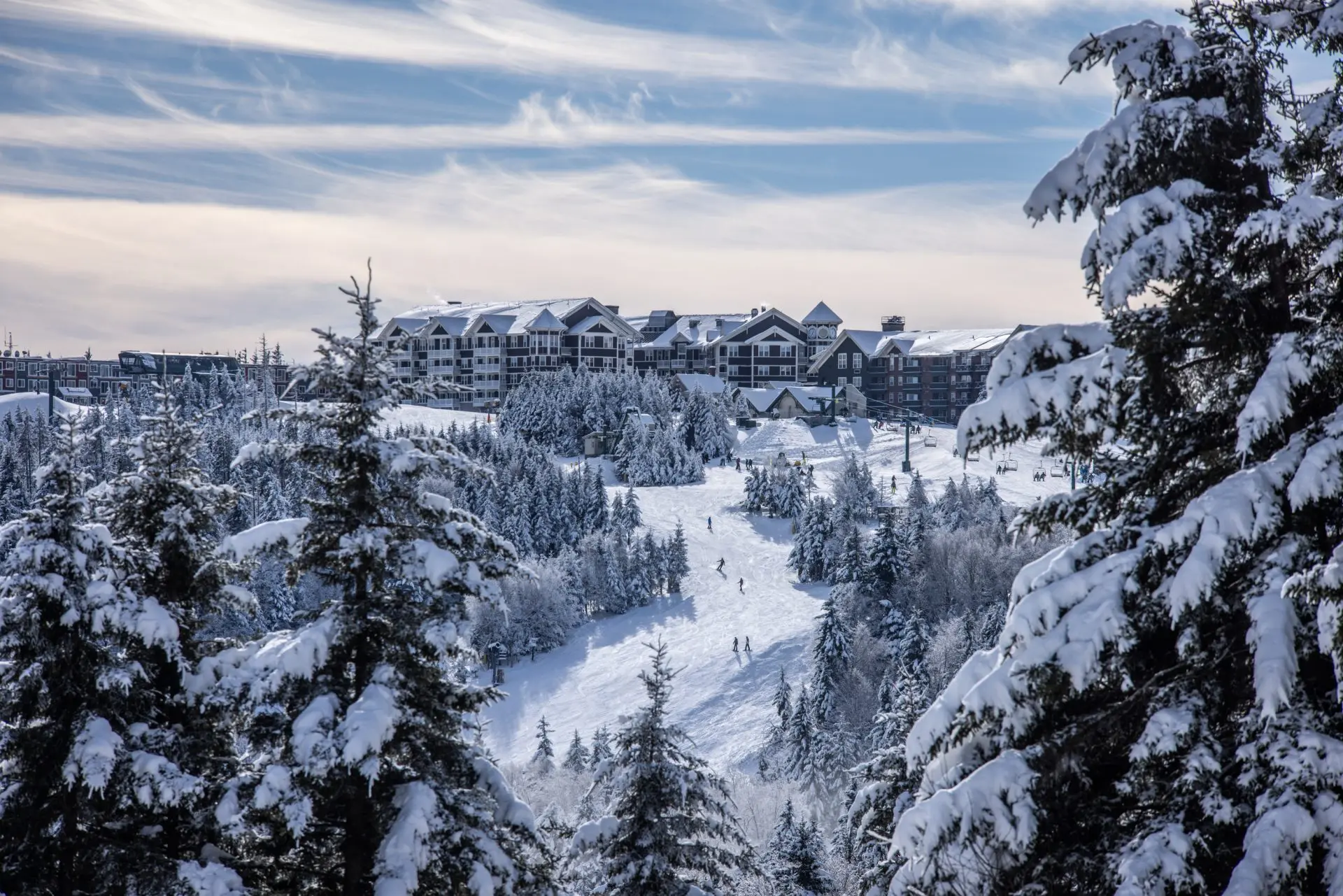 Snow-covered evergreens frame a ski slope leading to a large mountainside lodge under a partly cloudy sky.