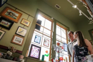 A man and woman smile while browsing a bright art gallery filled with framed paintings and stained glass artwork.