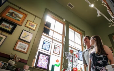 A man and woman smile while browsing a bright art gallery filled with framed paintings and stained glass artwork.