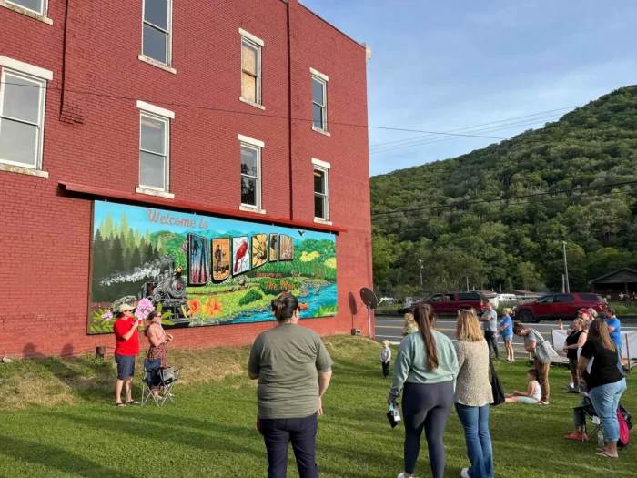 Crowd gathered on a lawn admiring a colorful "Welcome to Durbin" mural on a red brick building, with forested hills in the background.