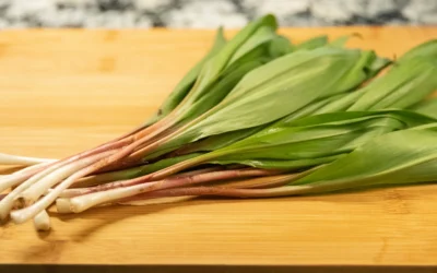 A close-up of fresh ramps on a wooden cutting board, showing the white bulbs, reddish-pink stems and wide green leaves, with a marble countertop visible in the background.