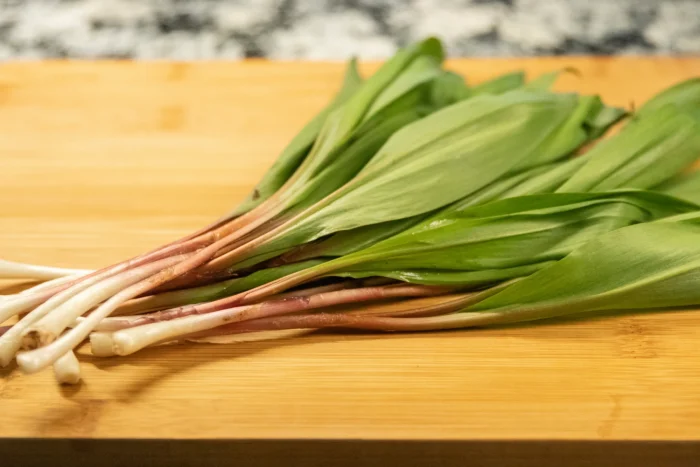 A close-up of fresh ramps on a wooden cutting board, showing the white bulbs, reddish-pink stems and wide green leaves, with a marble countertop visible in the background.
