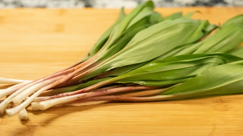 A close-up of fresh ramps on a wooden cutting board, showing the white bulbs, reddish-pink stems and wide green leaves, with a marble countertop visible in the background.