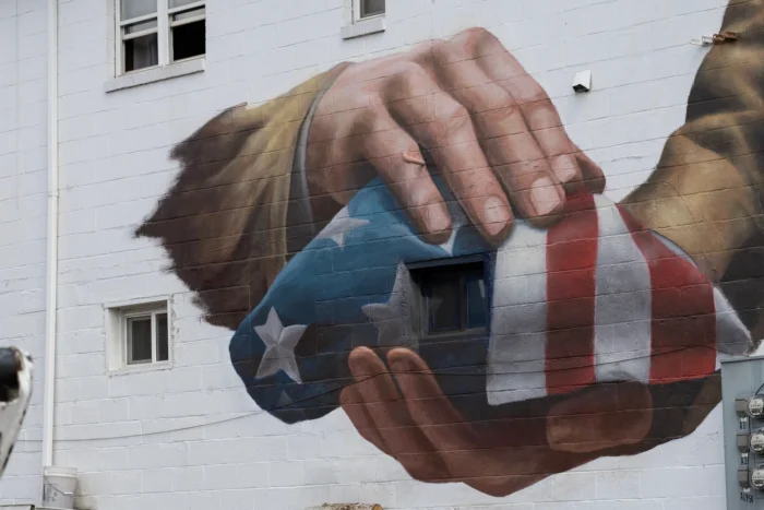 A large-scale mural of two hands cradling a folded American flag, painted on a white cinder block building.