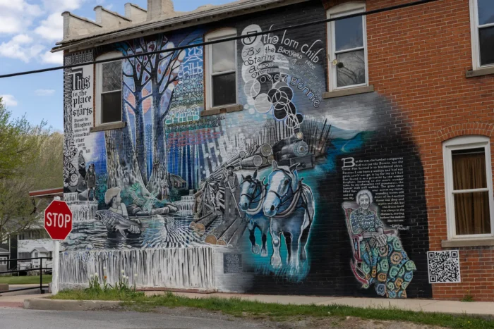 A detailed, text-filled mural depicting Appalachian history, including logging, horses and a river scene, on the side of a brick building.