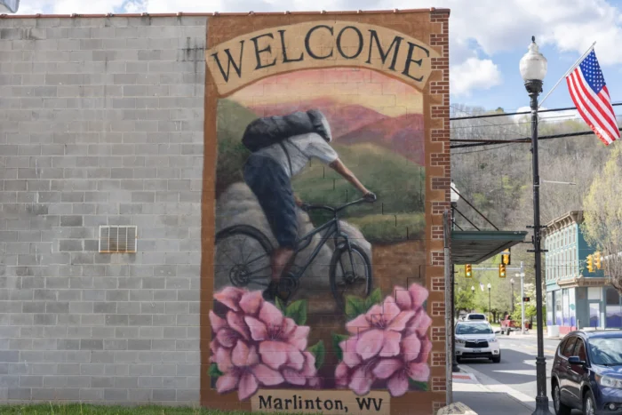 A "Welcome to Marlinton, WV" mural showing a mountain biker on a trail, framed by pink rhododendrons, on a brick building corner.