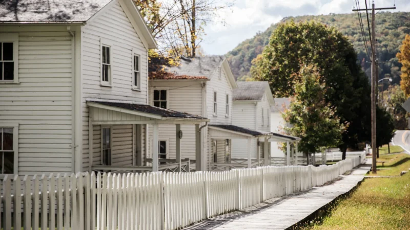 Row of white clapboard company houses with front porches and a white picket fence along a wooden boardwalk, with forested hills in the background.