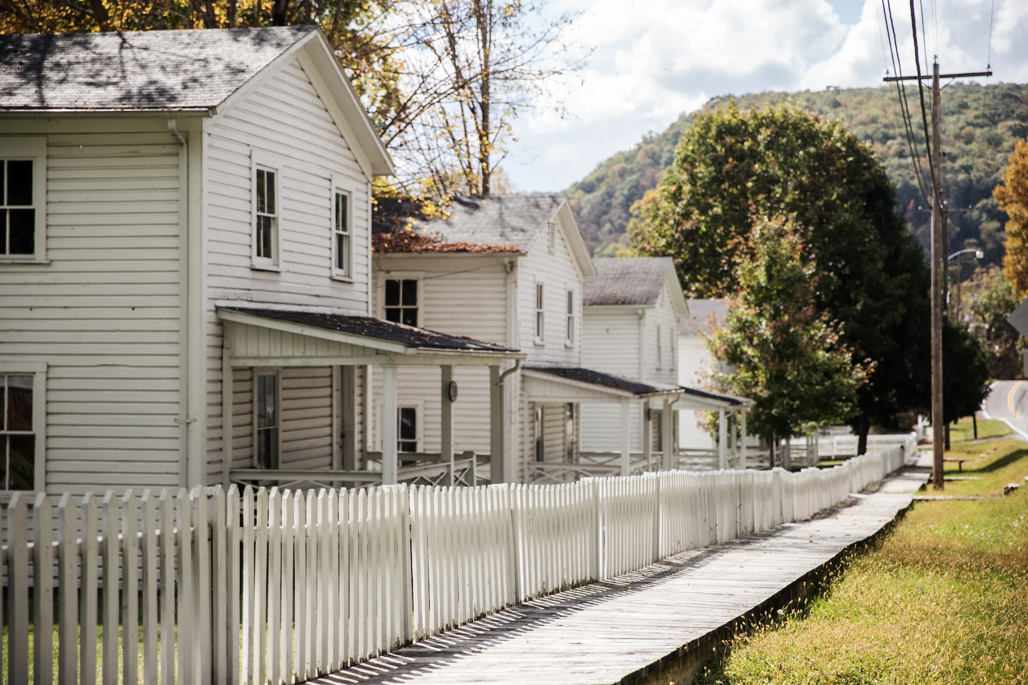 Row of white clapboard company houses with front porches and a white picket fence along a wooden boardwalk, with forested hills in the background.