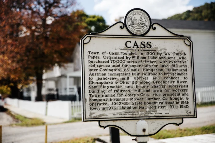 Historical marker for the town of Cass, West Virginia, with the state seal at the top, set against a backdrop of trees and a white building.