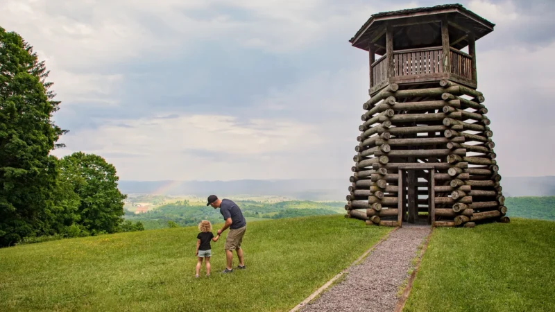 A father and his small child stand beside the lookout tower at Droop Mountain Battlefield State Park, looking out at the green valley below.